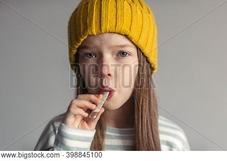 Portrait Of A Little Sad Unhappy Girl In A Hat, Runny Nose And Headache, Measures The Temperature Wi