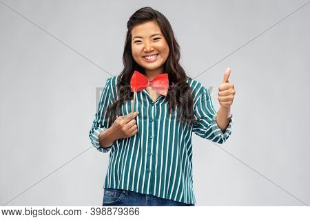 party props, photo booth and people concept - happy asian young woman with red paper bow showing thumbs up over grey background