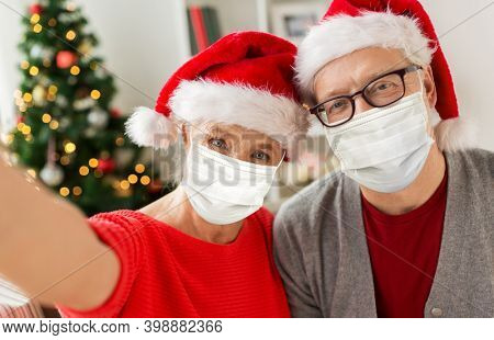 christmas, holidays and health concept - happy smiling senior couple wearing face protective medical mask for protection from virus disease in santa hats taking selfie at home