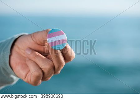 closeup of a young caucasian person outdoors showing a badge patterned with a transgender pride flag, in front of the ocean