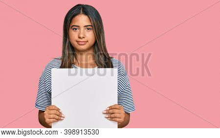 Young latin girl holding blank empty banner thinking attitude and sober expression looking self confident 