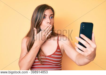 Beautiful young caucasian woman taking a selfie photo with smartphone covering mouth with hand, shocked and afraid for mistake. surprised expression 