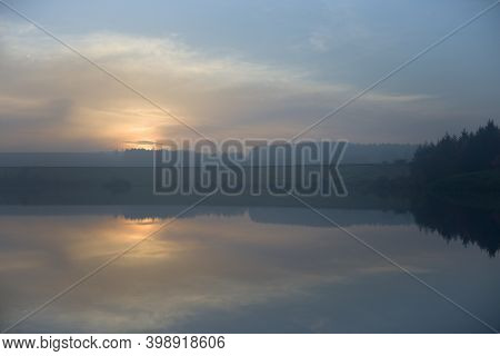 Dam Wall Acts As A Line Of Symmetry In This Reflected Landscape. Hazy Sunset Over The Calm Waters Of