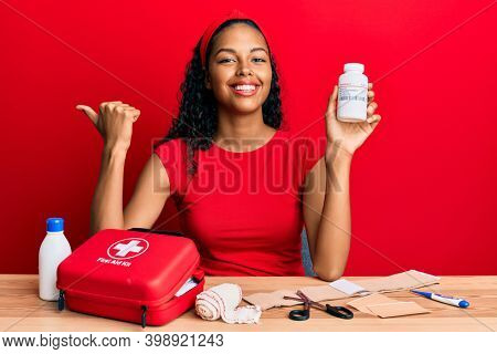 Young african american girl holding pills sitting on the table with first aid kit pointing thumb up to the side smiling happy with open mouth 