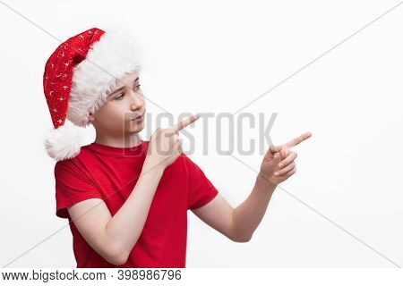 Portrait Of A Teenage Boy Wearing A Red Christmas Santa Claus Hat. Boy Points Finger On White Isolat