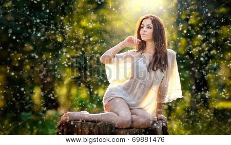 Young beautiful red hair woman wearing a transparent white blouse posing on a stump in forest