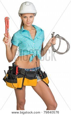 Pretty girl in helmet, shorts, shirt, tool belt with tools holding flexible hose and wrench