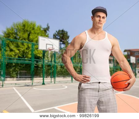Vertical shot of a young man with a blue cap holding a basketball and looking at the camera at an outdoor basketball court