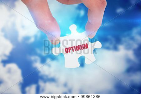 The word optimum and hand holding jigsaw piece against bright blue sky with clouds