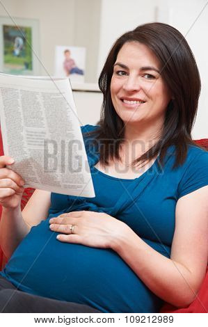 Pregnant Woman Sitting On Sofa Reading Magazine