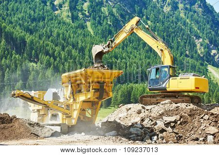 excavator loads the ground in the stone crusher machine during earthmoving works outdoors at mountains construction site