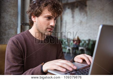 Concentrated serious handsome curly male in brown sweetshirt using laptop 