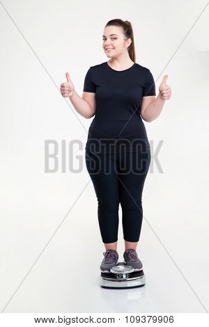 Full length portrait of a happy thick woman standing on weighing machine and showing thumbs up isolated on a white background