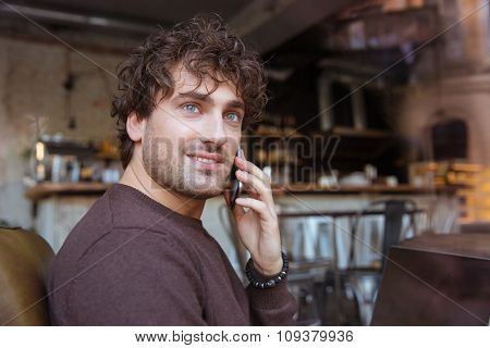 Handsome attractive smiling curly happy joyful content guy in brown sweetshirt sitting in cafe and talking on cellphone