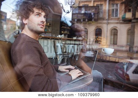 Smiling content curly young male in brown sweetshirt using laptop in cafe and dreaming