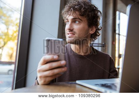 Thoughtful pensive handsome attractive young curly man in brown sweetshirt working with mobile phone and laptop 