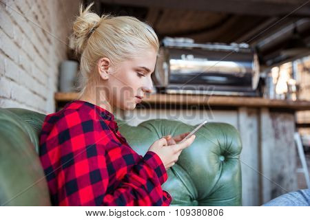 Young pretty attractive concentrated woman in checkred shirt sitting in cafe on green leather sofa and using mobile phone