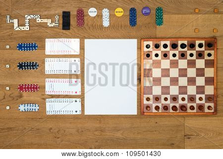 Top view of Board game items and gambling objects, such as a chess board and pieces, cards, chips, and dominoes neatly aligned on a wooden surface, seen from above, with a white area for your copy