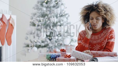 Young African woman wrapping gifts at Christmas sitting at a table in front of the Xmas tree chatting on her mobile phone  natural pre-occupied pose.