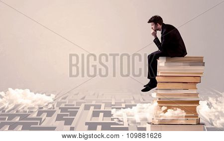 A serious businessman in suit sitting on a pile of giant books in front of a grey wall with clouds, labirynth