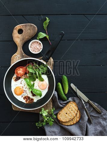 Pan of fried eggs, bacon, tomatoes with bread, mangold and cucumbers on rustic wooden serving board