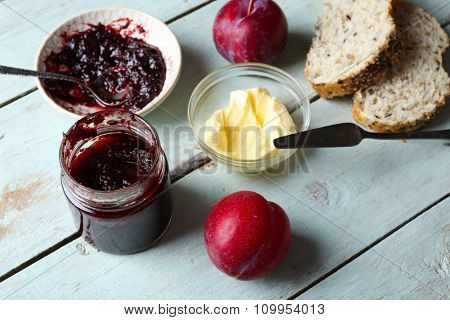 Tasty jam in the jar and bowl, butter, fresh bread and plums on blue wooden background