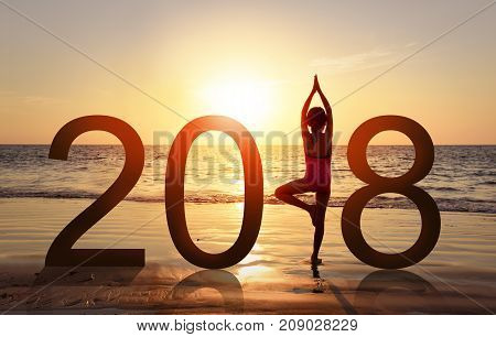 Happy new year card 2018. Silhouette of A girl doing Yoga vrikshasana tree pose on tropical beach with sunset sky background watching the sunset standing as a part of the Number 2018 sign.
