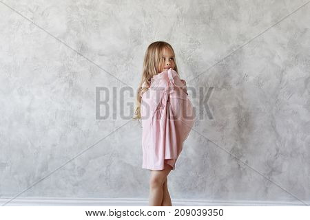 Adorable cute baby girl with long straight hair posing at blank grey wall wearing beautiful pink dress having shy look. Sweet female kid trying on fancy dress. Kids fashion and style concept