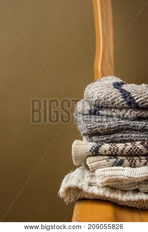 Stack of Handmade Warm Knitted Socks Scarfs Mittens Sweaters From Rough Wool Yarn Brown Beige Grey on Vintage Chair Golden Olive Color Wall Background. Eco Fashion Kinfolk Style. Interior