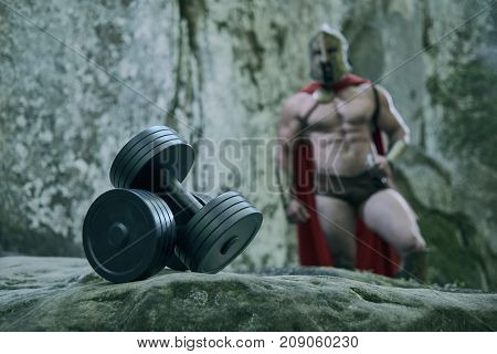 Selective focus on heavy dumbbells strong muscular Spartan warrior on the background showing off his muscles.