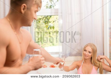 Young muscular man giving a glass of champagne to pretty blonde in bicini sitting in a foam bath shot in sauna
