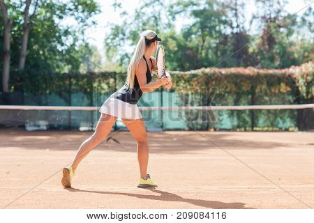 Full body photo of sexy young lady tennis player in action in a tennis court indoor. Photo of the woman is made from the back. Blonde tennis player dressed white skirt, black T-shirt and black cap