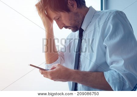Businessman standing next to office window and using smartphone. Business communication over sms messages or mobile apps. Business person received bad news.