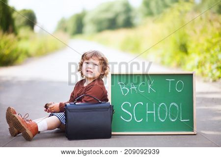 Cute toddler girl wearing school uniform with back to school blackboard wautung gor a school bus. Fall outdoors education concept sunny autumn day. Early education little genius wunderkind concept.