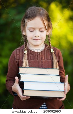 Adorable happy little preschooler girl with pigtails ready back to school holding pile of textbooks manuals. Wearing school uniform. Warm september fall day. Smart clever intellegent little girl.