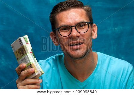 Portrait of young stylish man wearing eyeglasses with heaps of money. Cheerful male adult posing with cash on blue background.