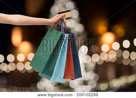 sale, holidays, consumerism and people concept - close up of hand holding shopping bags and credit card over christmas tree lights background