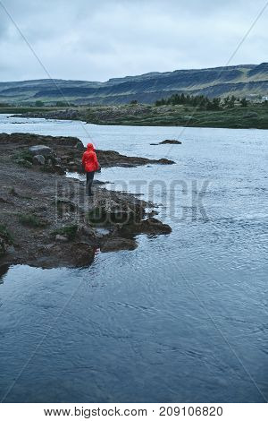 a woman tourist in waterproof clothes and red hat stands on the Bank of the mountain river with small waterfall. Glanni waterfall, Iceland
