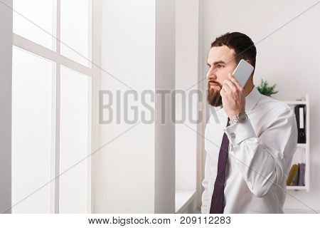 Important call. Young businessman in official colothes has cell phone talk in modern white office interior, copy space.