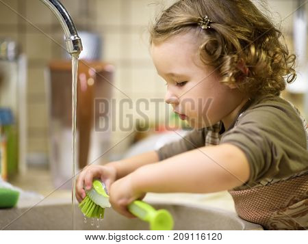 Pretty two-year girl washes the dishes in kitchen. Little child uses a brush for washing of ware.