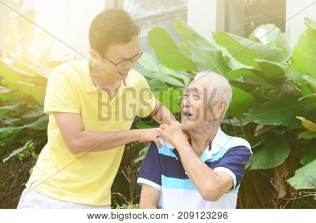 Asian old man shoulder pain son massaging father shoulder. Chinese family senior retiree outdoors living lifestyle in the garden.