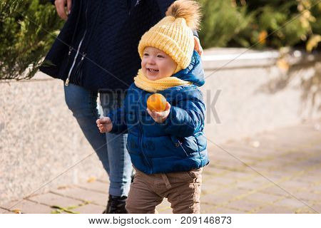 happy little boy in the rays of the sun in autumn, in the leaves and a yellow cute cap holding clementine