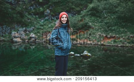 a woman tourist in waterproof clothes and red hat stands on the Bank of the mountain lake with turquoise water. Glanni waterfall, Iceland
