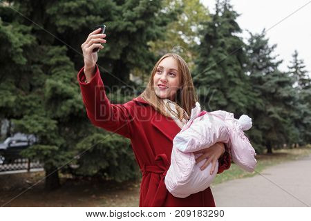 Happy young mommy spend time with a little baby in the park outdoors. A pretty woman taking selfie with baby on the nature background. Family concept.