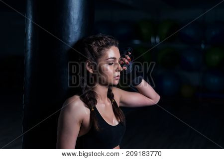 Upset young fighter boxer girl wearing boxing gloves, standing in dark gym near punching bag after loosing fight. She coveres her face with hand.