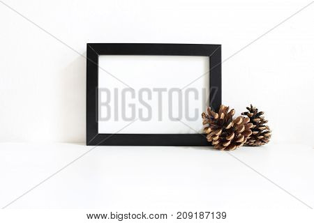 Black blank wooden frame mockup with pine cones lying on the white table. Poster product design. Styled stock feminine photography, home decor. Christmas winter concept.