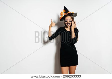 Halloween witch in a terrible suit and hat shows thumbs up and laughs. The girl was going to the Halloween party.