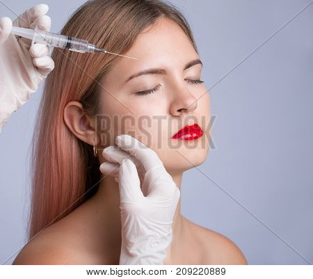Beautiful young woman with red lips gets cosmetic injection, isolated over greye background. Doctor is making an injection in face. Beauty Treatment.