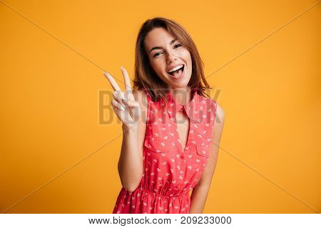 Close-up portrait of funny brunette girl showing peace gesture, looking at camera with open mouth, isolated on tellow background