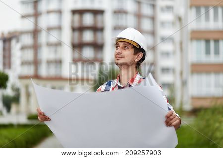 Young male constructionist smiling looking away holding blueprint of a building under construction future development real estate business industry businessman concept